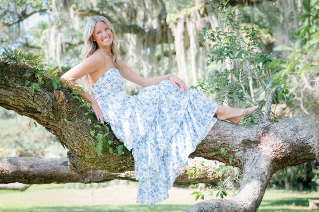 High school senior sitting on a tree branch in a flowing dress, striking a playful pose during a portrait session.