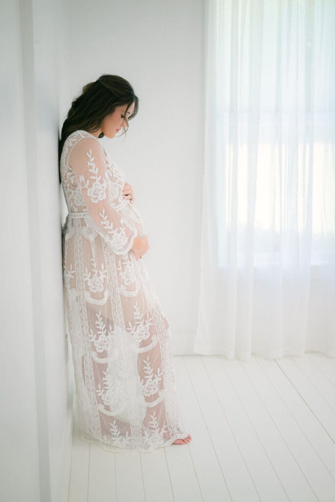 Maternity photo of a woman in white lace dress, leaning on wall and looking down at her baby bump