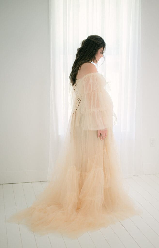 Full length maternity portrait of a woman standing in front of a bright window