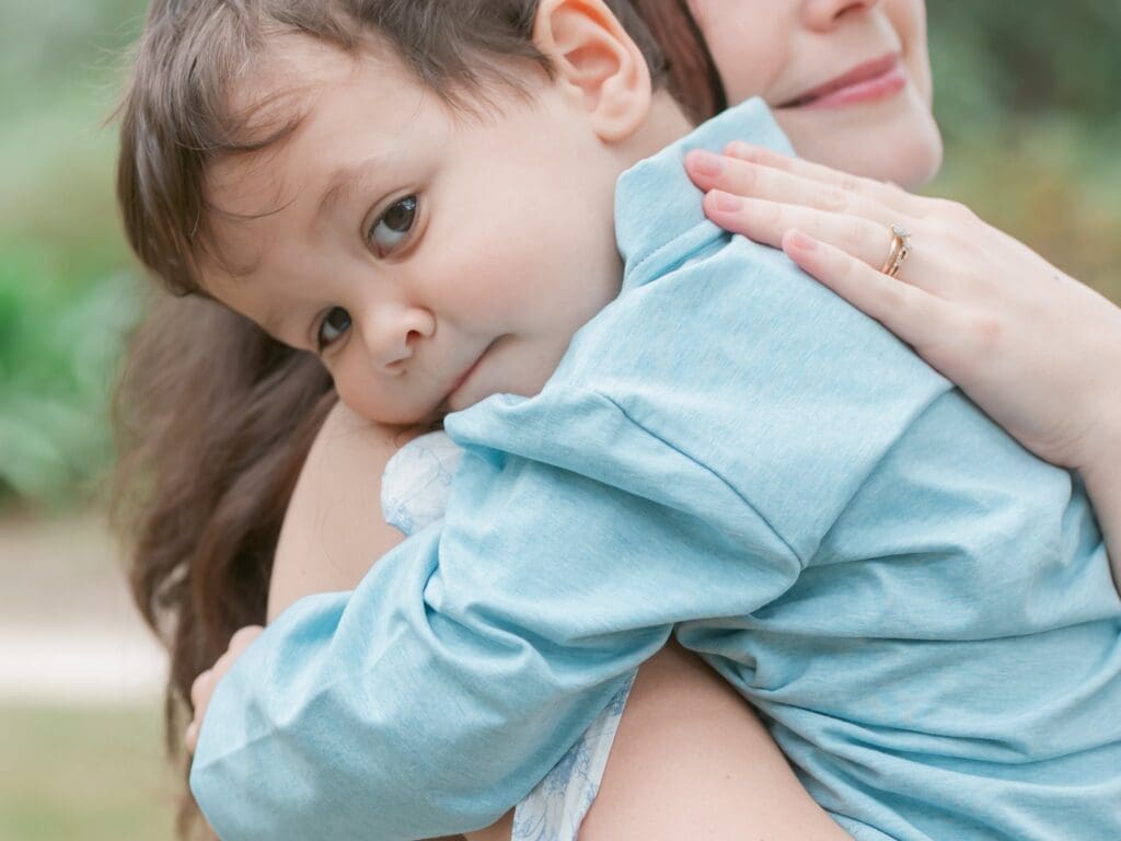 Close-up outdoor portrait of a mother gently holding her toddler son as he rests his head on her shoulder, captured during a natural motherhood photography session in Tallahassee, Florida