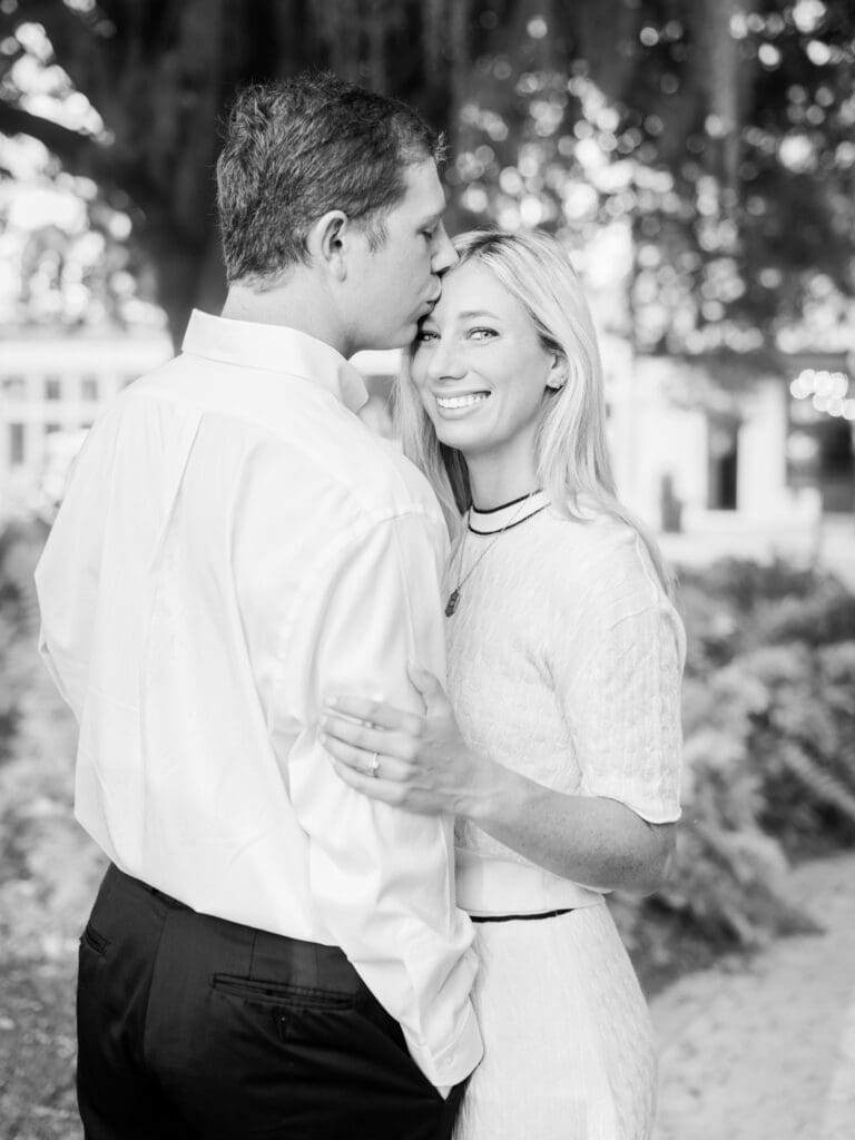 Black and white photo of man kissing his fiancée’s forehead while she smiles at the camera during proposal