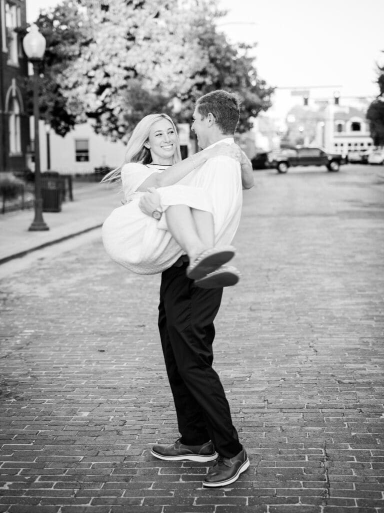 Black and white of groom swinging his fiancée in the air while they laugh after proposal