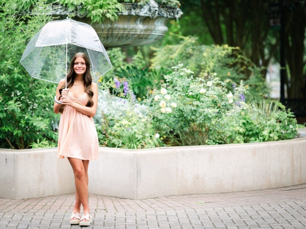High school senior standing in front of a flower-filled fountain in the misting rain while holding a clear bubble umbrella during a senior portrait session in Tallahassee, Florida.