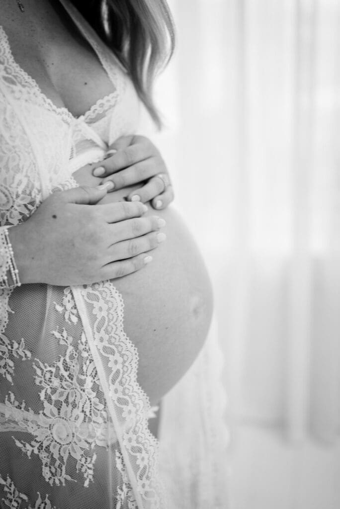 Pregnant woman standing near a window holding her belly in a timeless maternity portrait.